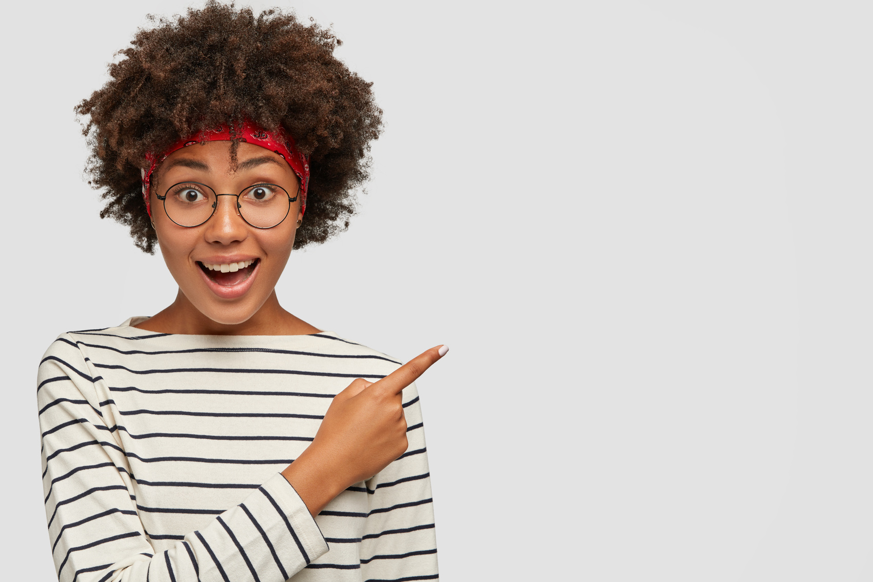 Studio shot of positive dark skinned girl wears round transparent glasses, striped white sweater, indicates with index finger at upper right corner, shows free space for advert. Promotion concept